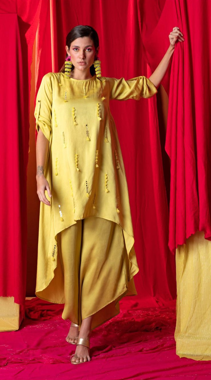 Image of Our Yellow Tunic Twin Set features a eye-catching high-low hemline adorned with mirror and multi-colored tassels. The set is perfectly complemented with an asymmetric palazzo, making it an ideal pick for summer events.  From savoirfashions.com